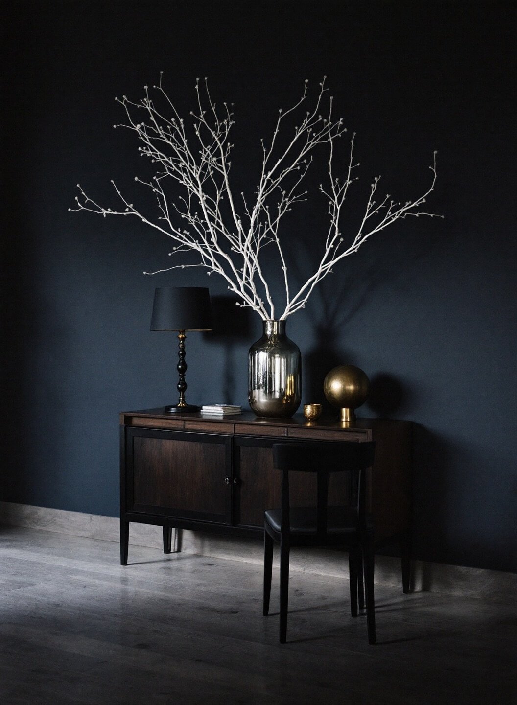 Dining room sideboard decor at dusk featuring dark walnut credenza, black metal table lamp, mercury glass vase with white branches, and small brass object against a deep navy wall under warm ambient light