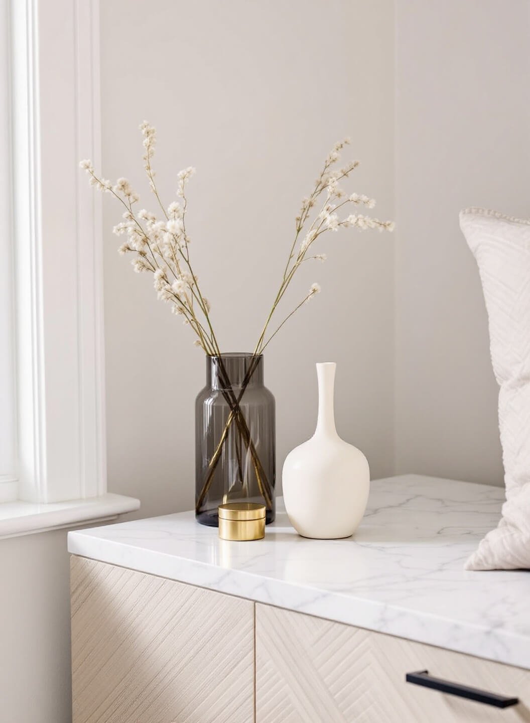 Contemporary bedroom dresser with smoky glass vase, cream ceramic vessel, and brass trinket box bathed in soft morning light from a nearby window