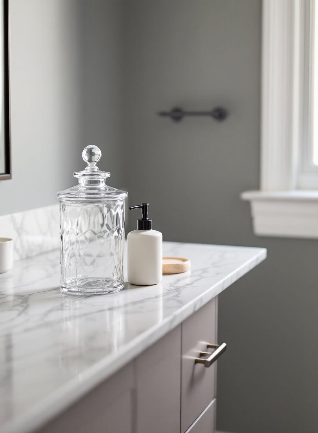 Close-up of a master bathroom vanity at dawn with a glass apothecary jar, ceramic soap dispenser, and natural stone dish on a white marble counter, illuminated by diffused window light from the right