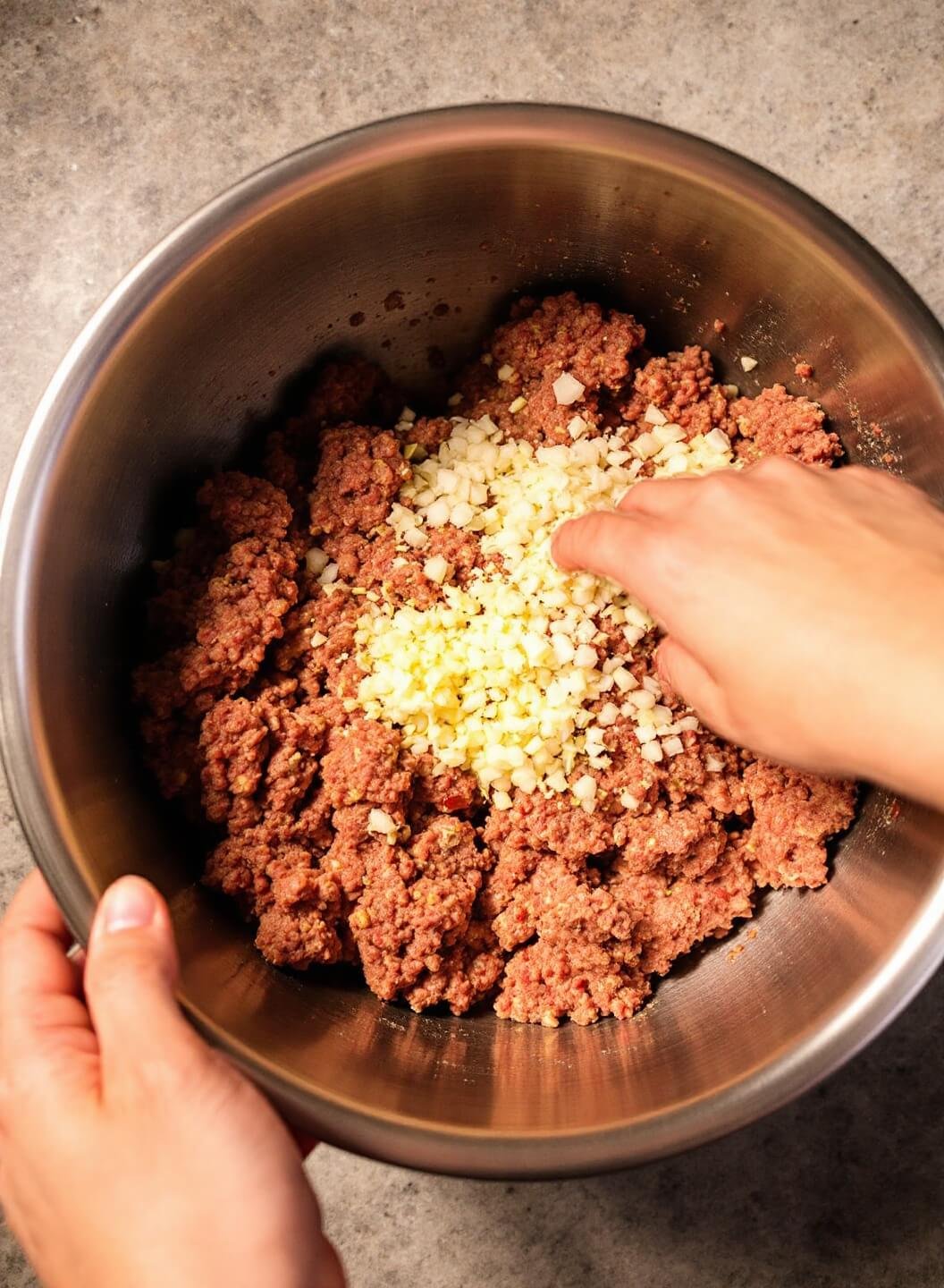 Hands combining minced garlic, onion and meat in a stainless steel bowl under warm kitchen lighting