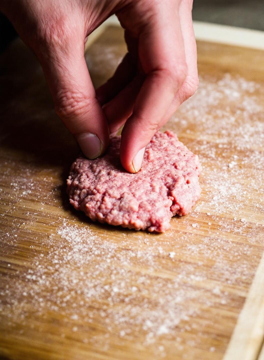 Hands gently shaping ground meat into perfect patties on a wooden cutting board, creating a dimple in the center with a light dusting of spices visible.