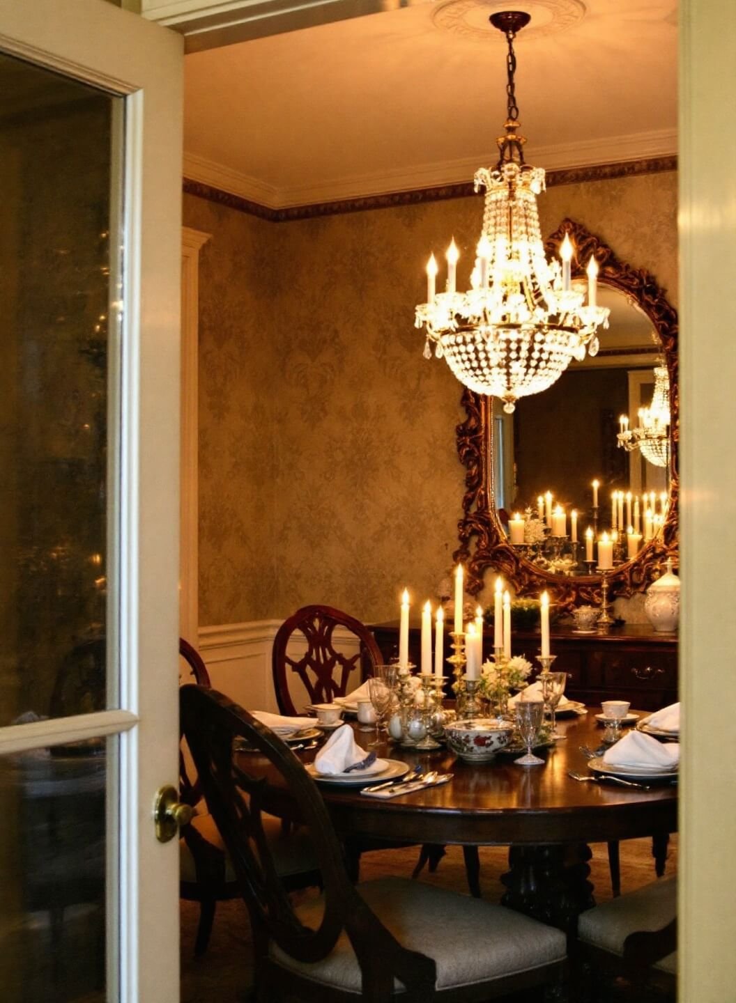 Traditional dining room at dusk, lit by a crystal chandelier's warm light, Louis XVI chairs surround a mahogany table set with fine china and silver, reflected in a large gilded mirror on a damask-papered wall, captured from a low angle at the doorway.