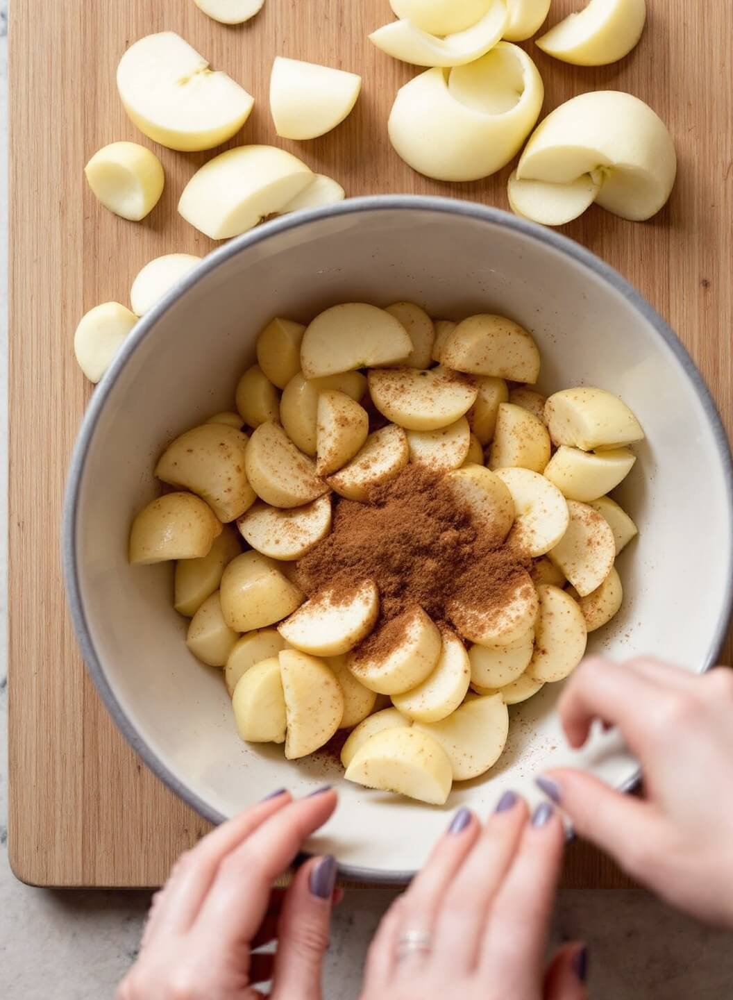 Hands peeling and slicing apples onto a wooden cutting board, with a bowl of apple slices mixed with cinnamon and sugar nearby.
