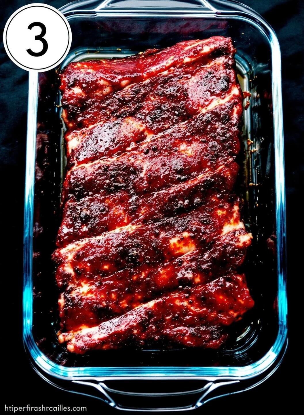 Marinated deep red glazed ribs in a clear glass container in the refrigerator, showcasing layers of marinade coating and aromatic ingredients