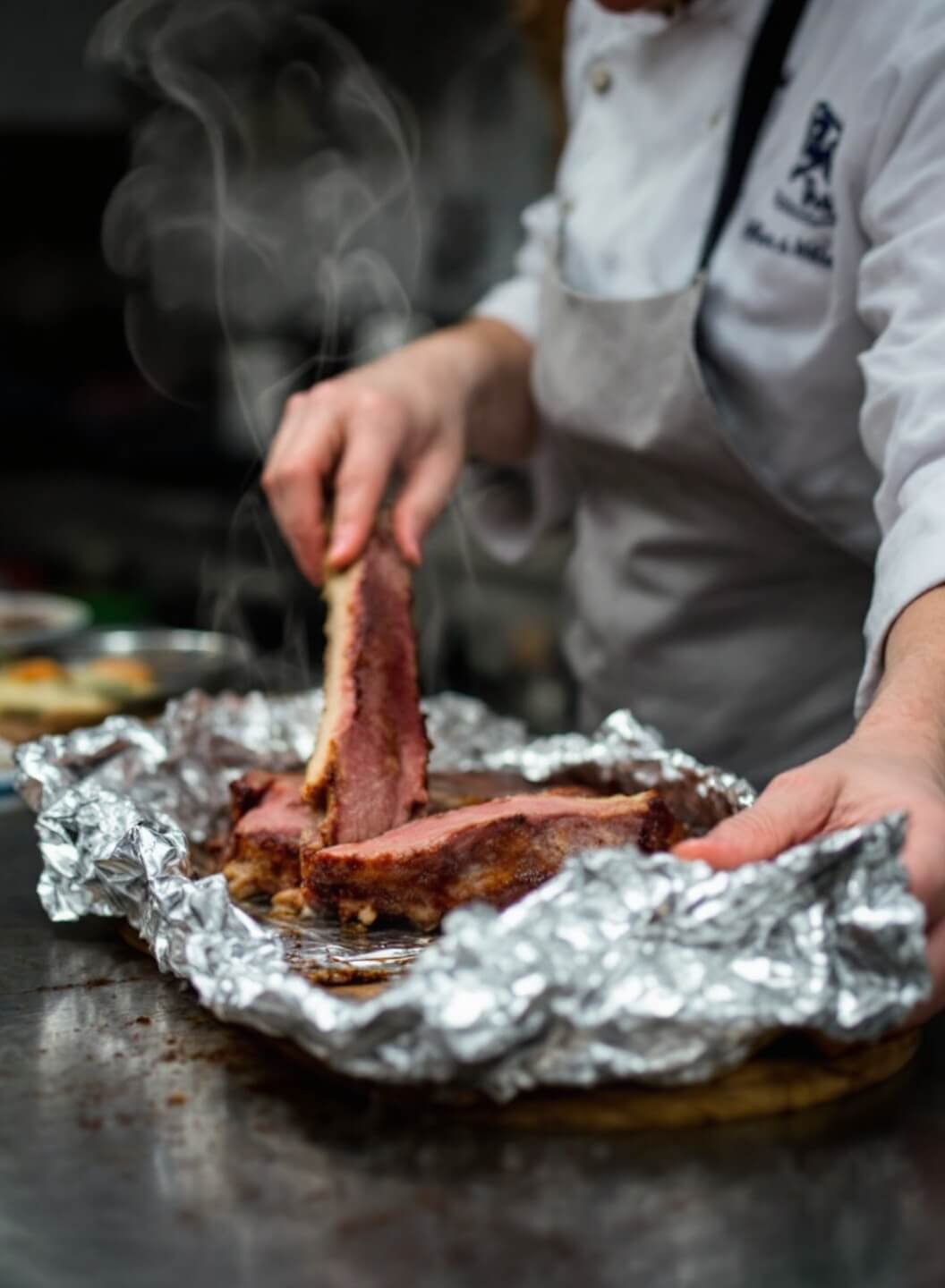 Chef carefully unwrapping foil from steaming ribs, revealing tender meat pulling from the bone in a professional kitchen