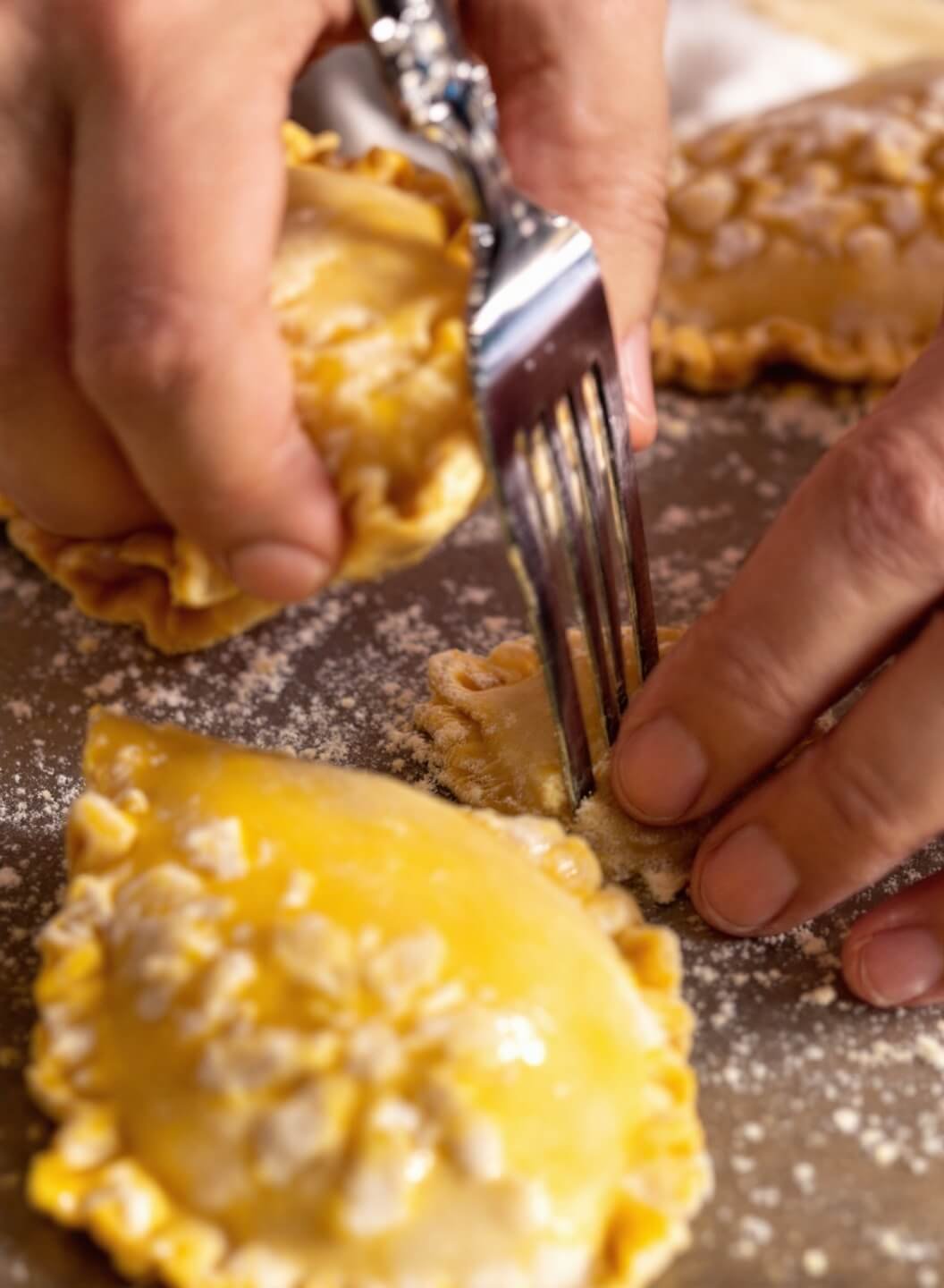 Hands crimping edges of an empanada with a fork, egg wash glistening under warm kitchen light with flour scattered in the background.