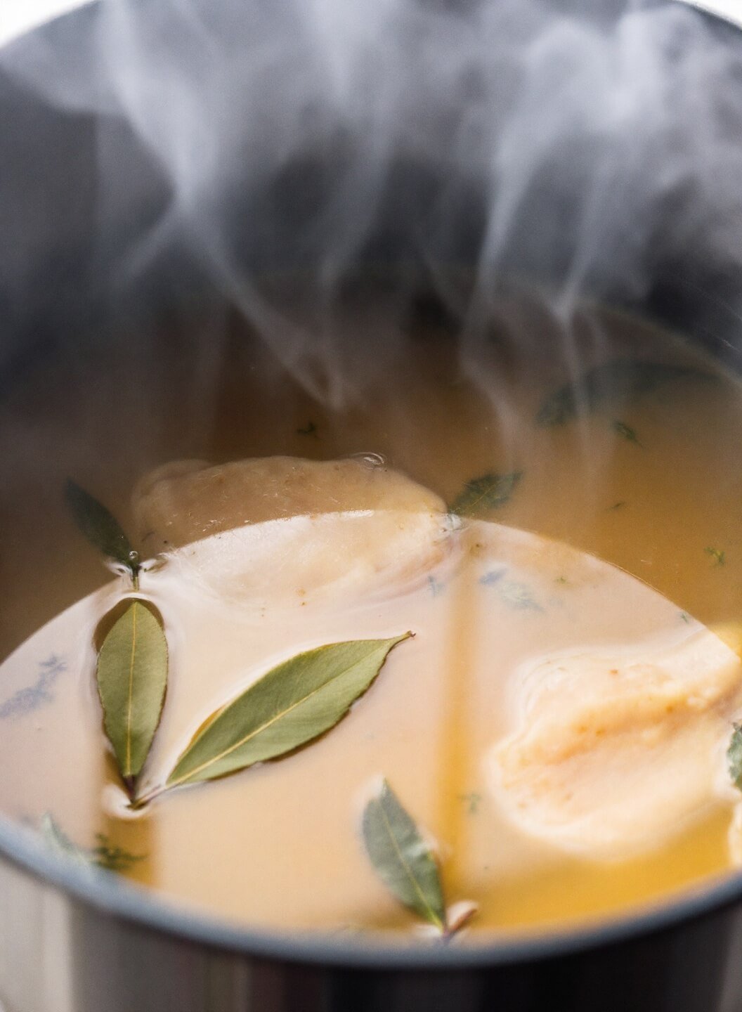 Steam rising from simmering pot of golden chicken soup with visible pieces, bay leaves, and herbs