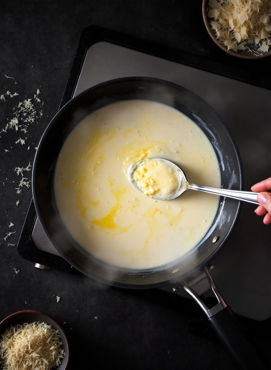 Overhead view of cream sauce being prepared in a skillet with melted butter, minced garlic, steam and grated Parmesan under dim restaurant-style lighting