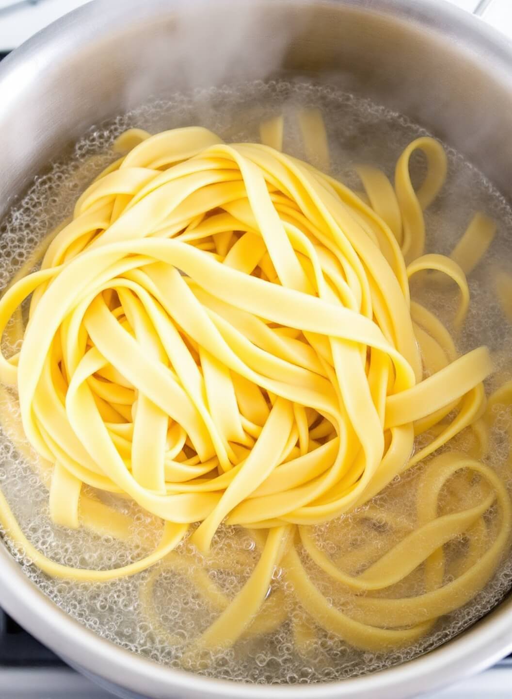 Fettuccine pasta being stirred in a large boiling pot, under bright kitchen lights