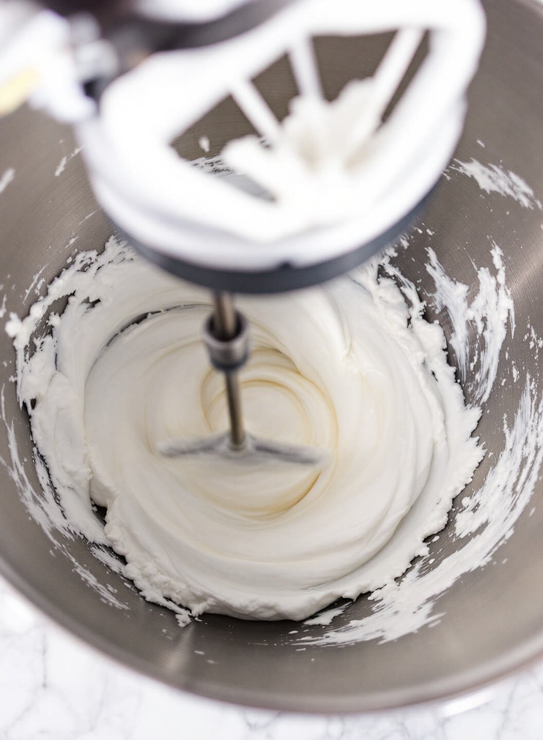 Whipped egg whites forming soft peaks in a stainless steel bowl under kitchen lighting, with mixer in action.