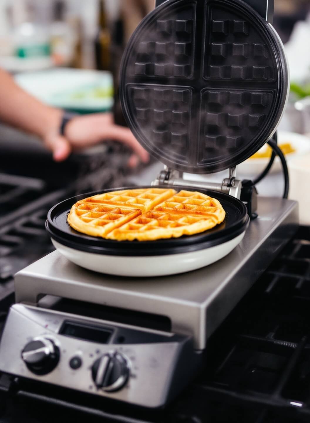 Closeup of a golden-brown waffle rising from a steaming waffle iron in a professional kitchen.