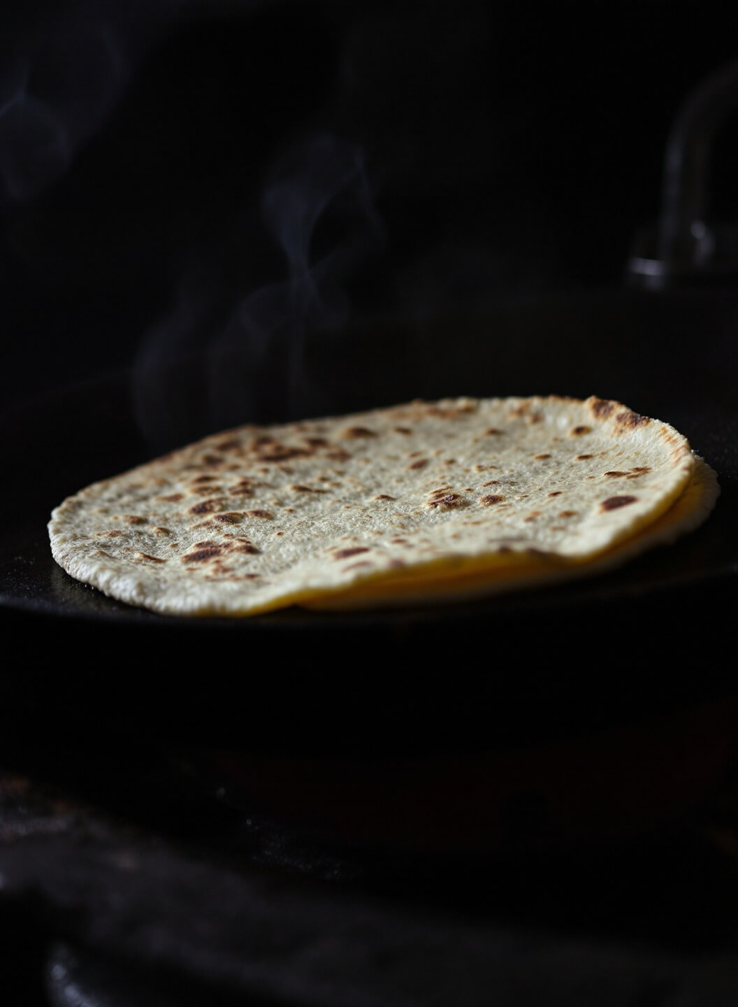 Corn tortillas warming on rustic comal with visible char marks and rising smoke in moody lighting