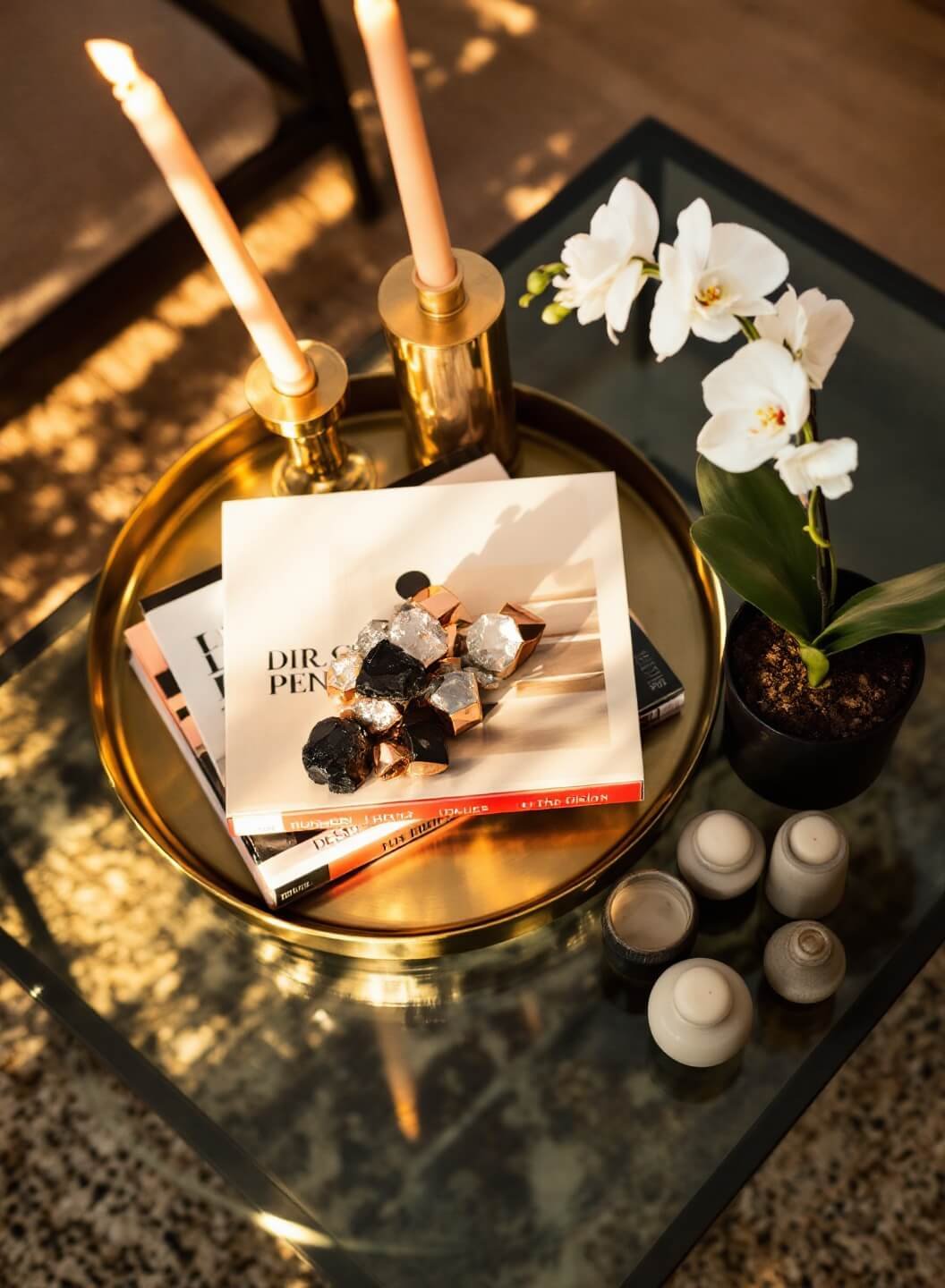 Overhead shot of square glass coffee table with vintage brass tray, crystal clusters, design magazines, white orchid in black pot, and cream ceramic objects in a contemporary apartment during golden hour
