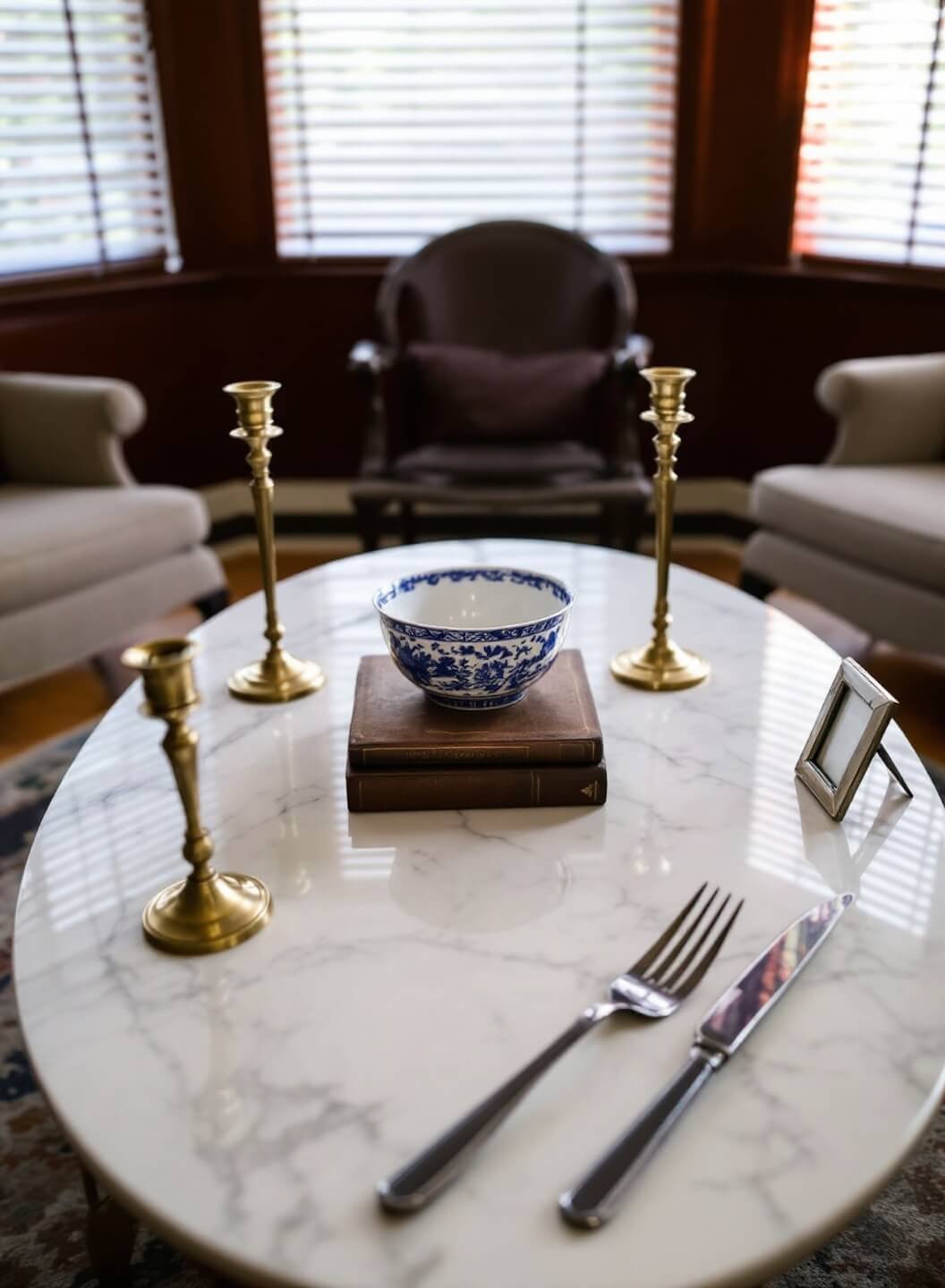 Marble coffee table with vintage books, brass candlesticks, and chinoiserie bowl in a traditional sitting room bathed in soft afternoon light from venetian blinds