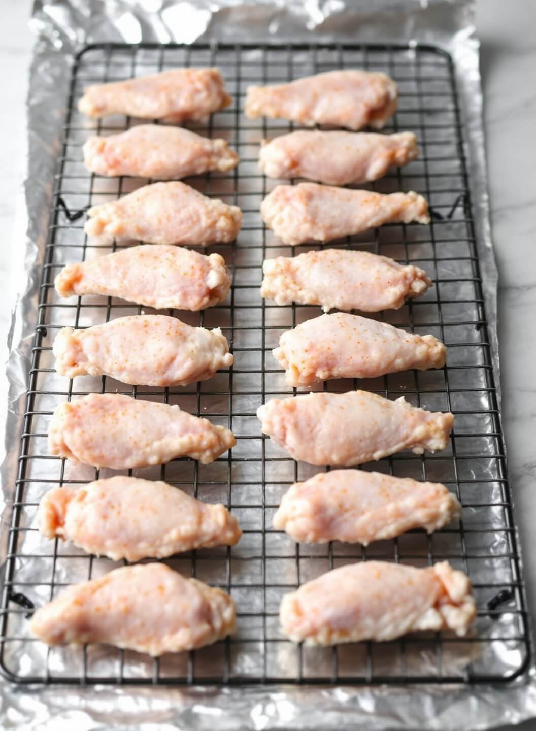 Seasoned raw chicken wings arranged precisely on a wire rack over a foil-lined baking sheet, highlighted with soft side lighting