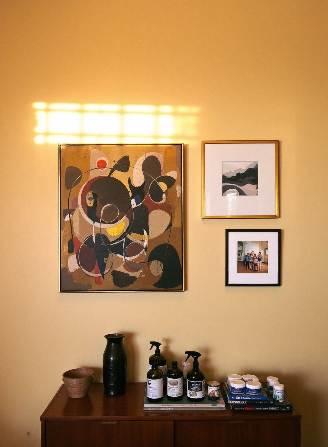 Mid-century modern den with beige walls, warm golden hour light illuminating a gallery of abstract art and family photos in brass frames, placed above a teak credenza.