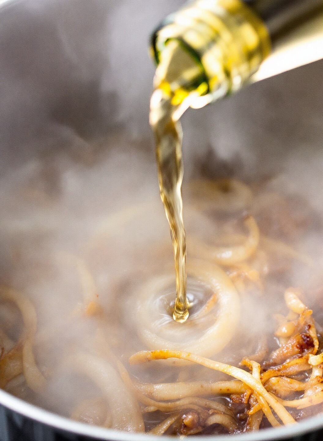White wine being poured into a steaming pot with caramelized onions, creating a sizzling effect with visible steam and light-catching droplets. Brown fond on pot sides.