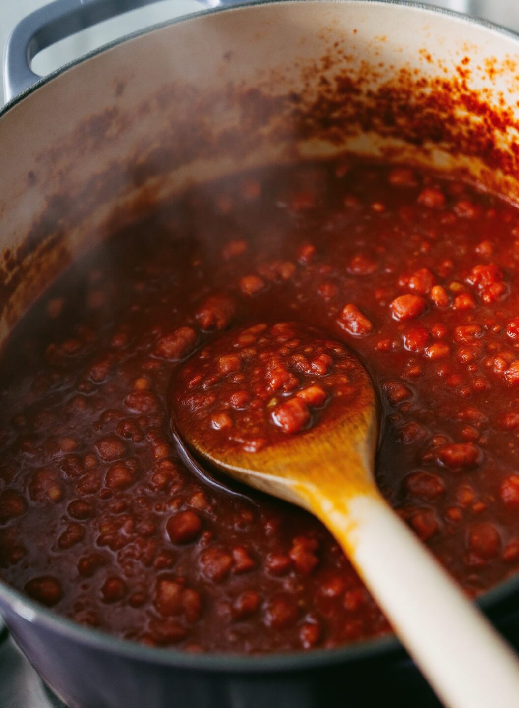 Simmering red meat sauce in a Dutch oven with wooden spoon and visible aromatics under warm kitchen lighting