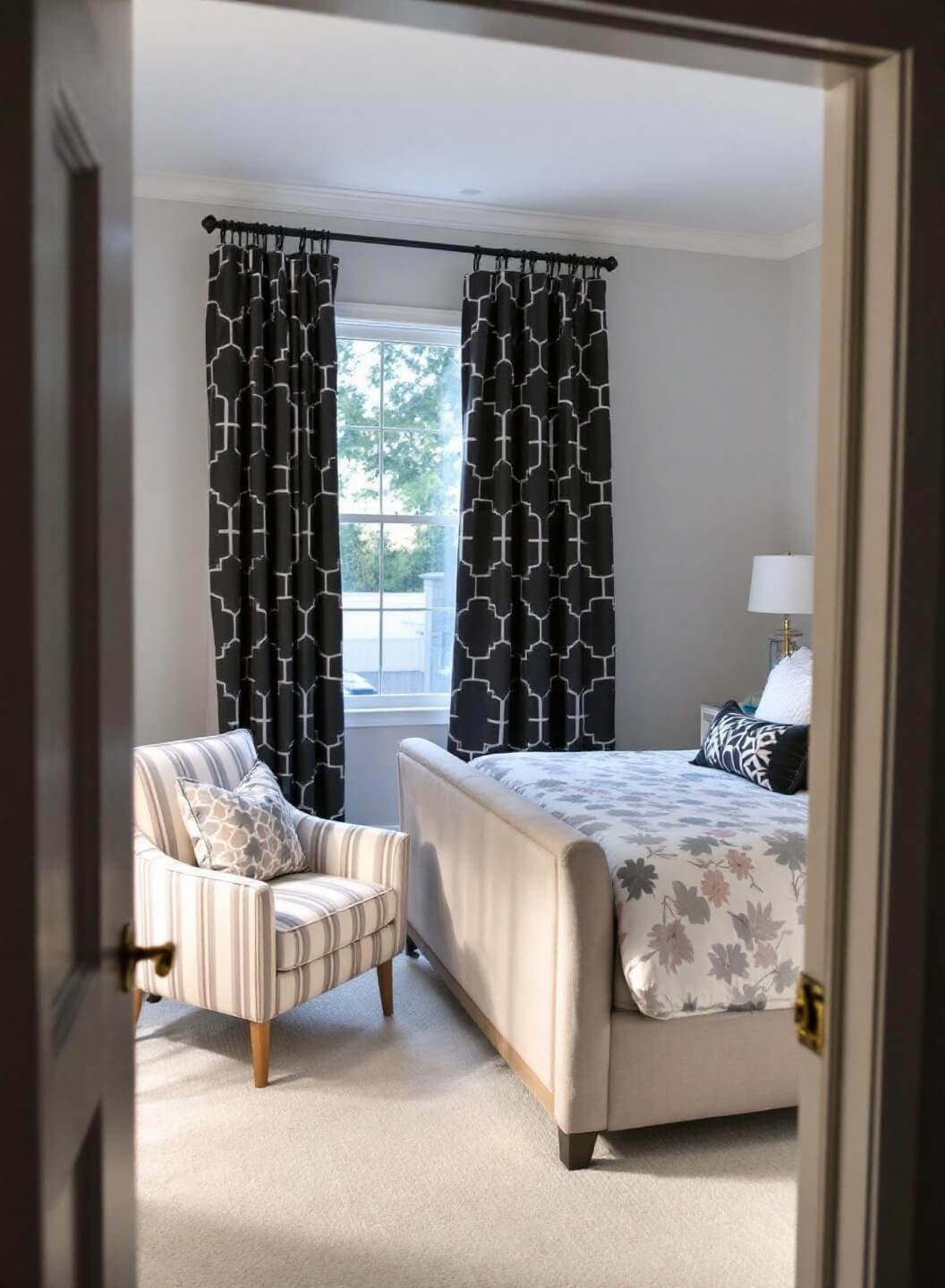 Modern master bedroom at dawn with blush and gray bedding, floor-to-ceiling windows with charcoal geometric print curtains, and a coordinating striped accent chair.