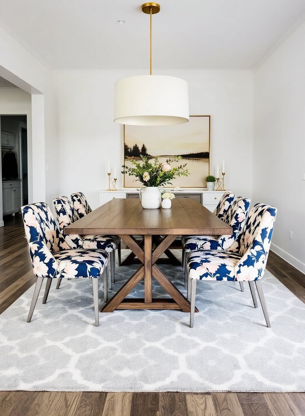Contemporary dining room with walnut table, geometric area rug, floral upholstered chairs in navy, blush and cream, brass candlesticks, white walls, and a dramatic overhead pendant, under moody evening lighting.