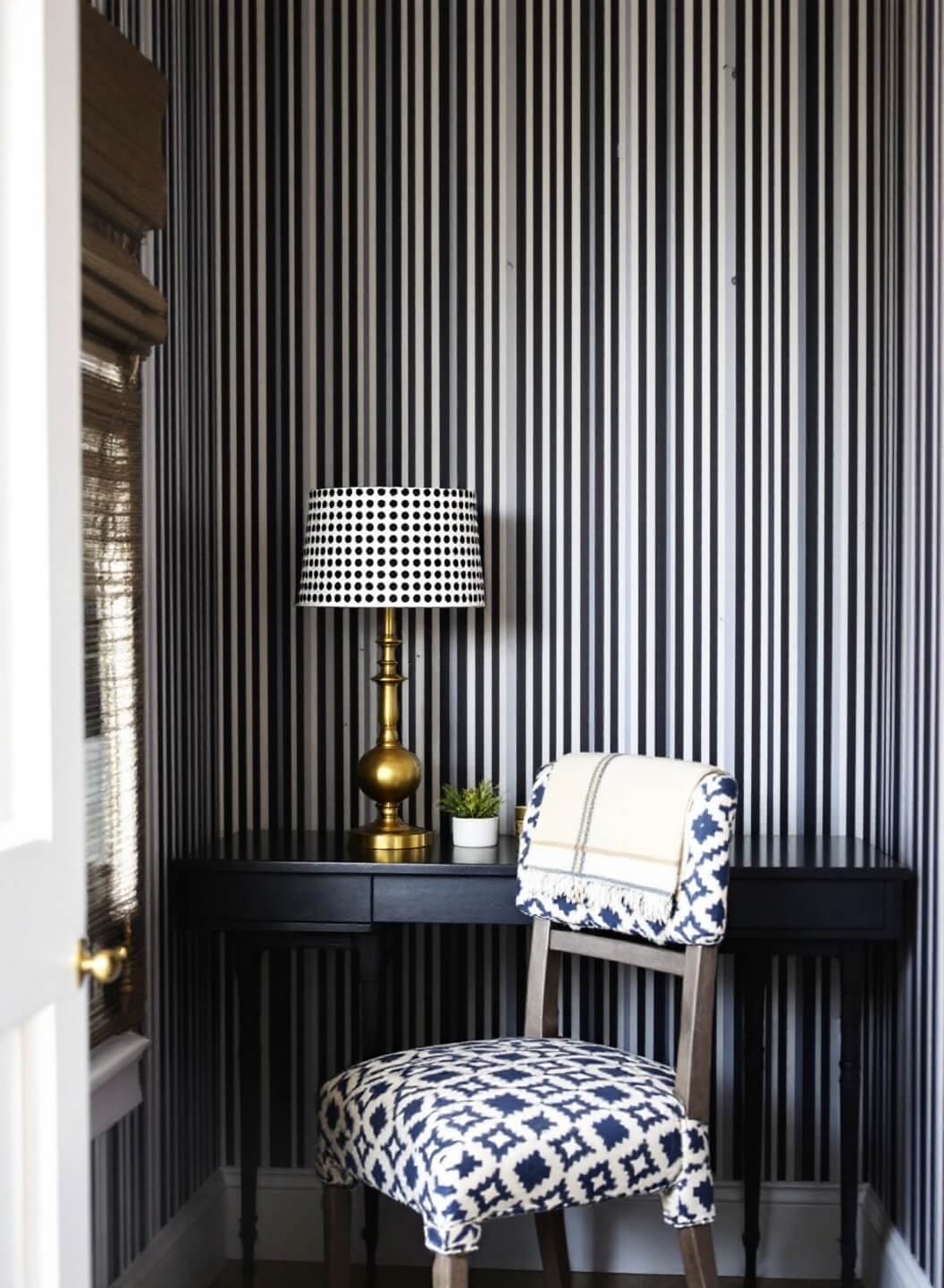 Midday shot of a compact 8x8 ft home office nook with vertical striped charcoal and white wallpaper, a navy and cream diamond-patterned desk chair, and a small brass lamp with dotted shade, enhanced by strategic side lighting.