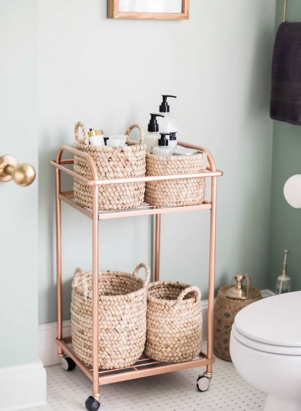 Rose gold rolling cart with woven baskets containing bathroom essentials, nestled between white toilet and seafoam green wall in softly lit, texture-rich setting