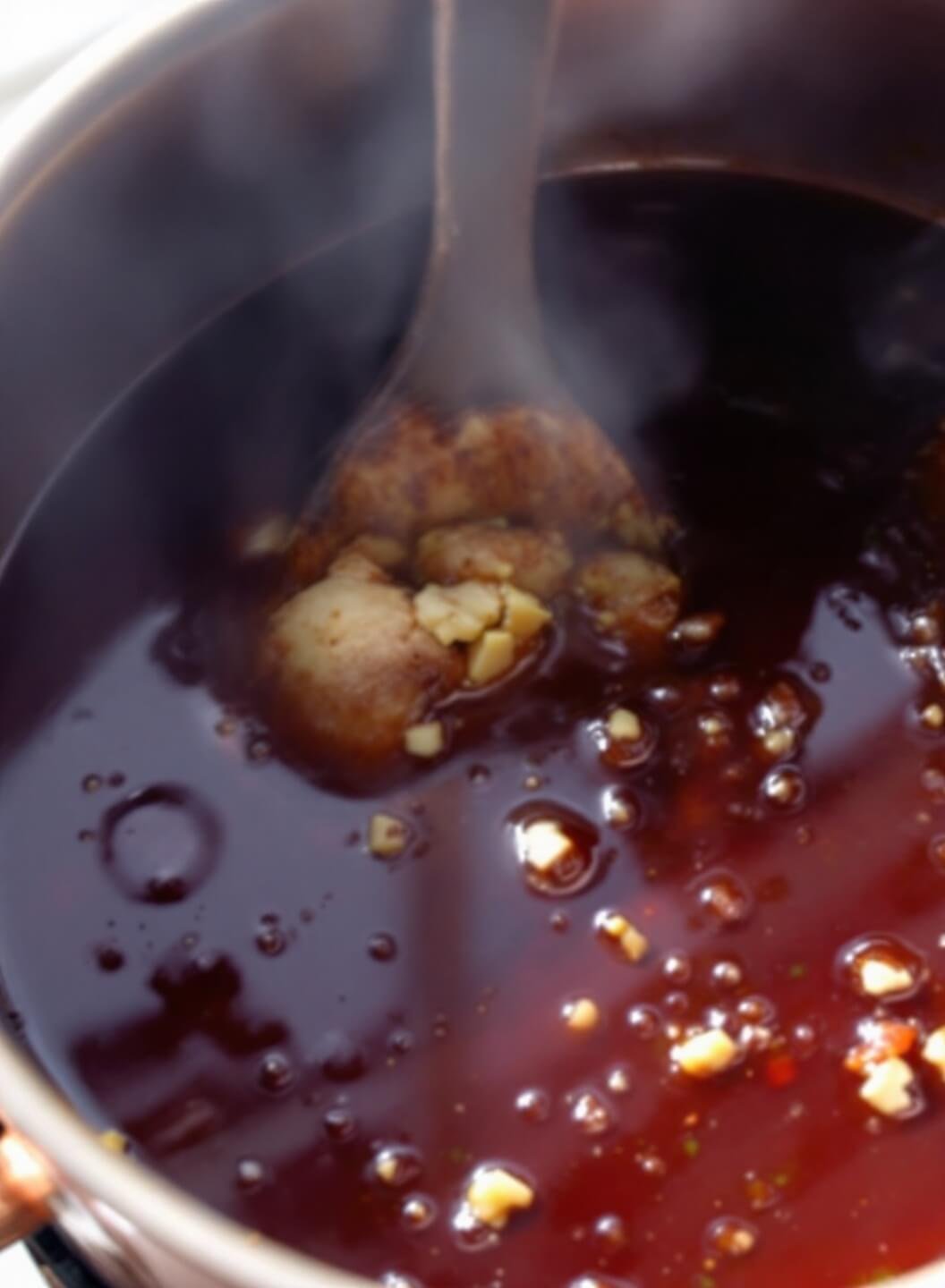 Bubbling BBQ sauce in copper saucepan with wooden spoon, fresh ginger and garlic in soft window light