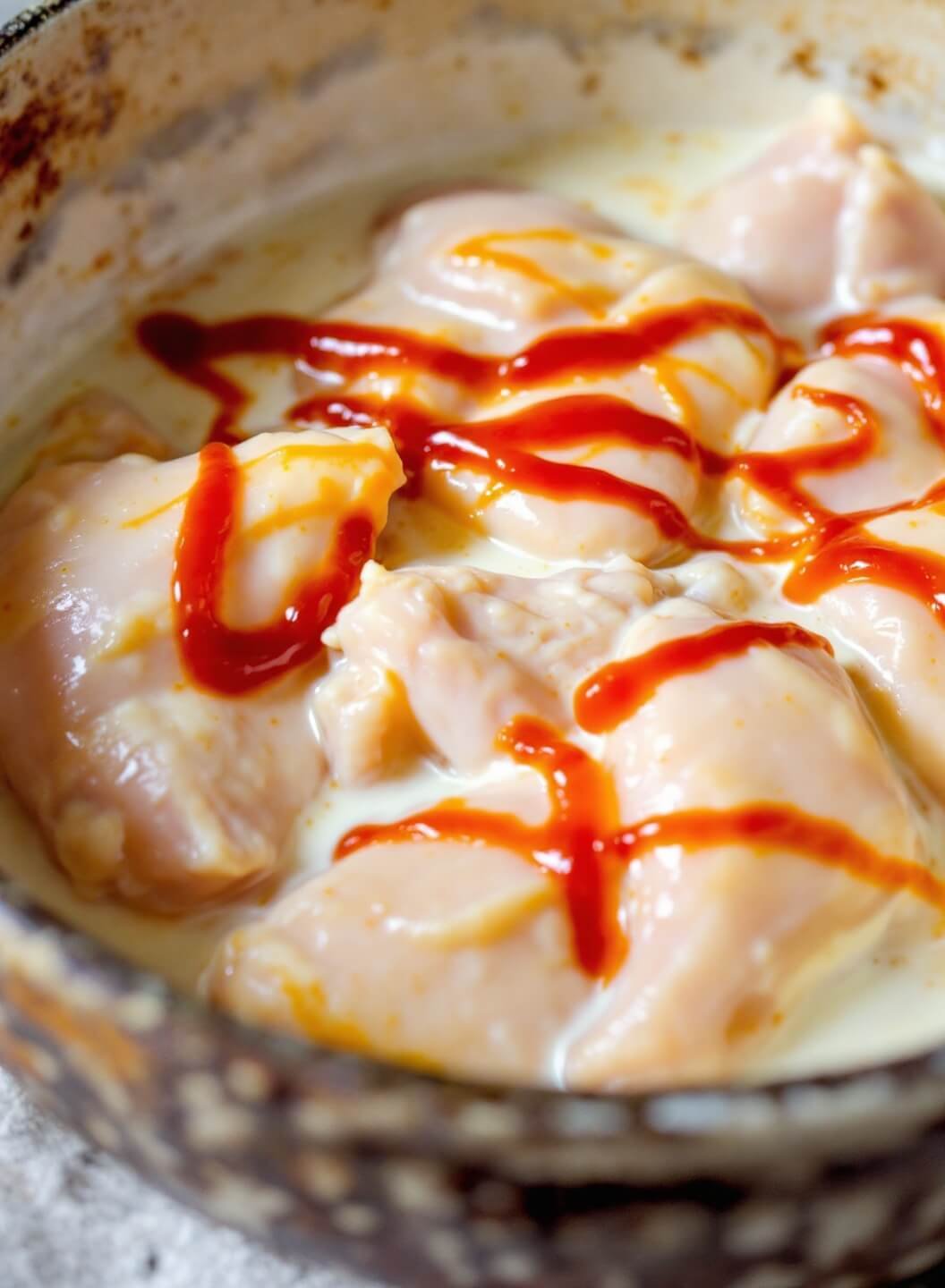 Raw chicken soaking in buttermilk marinade with hot sauce swirls in a stoneware bowl