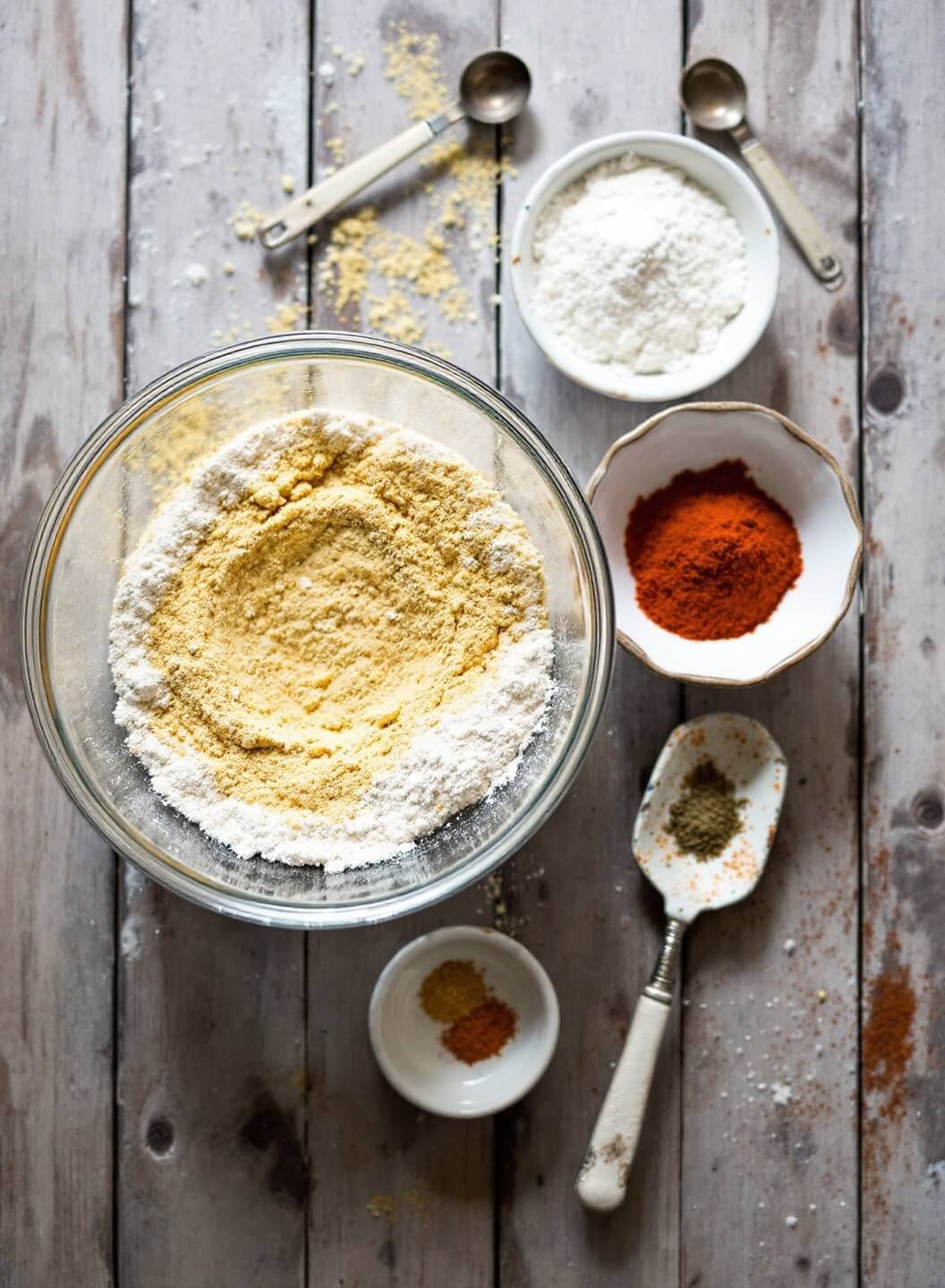 Overhead view of seasoned flour preparation on a rustic wooden table with bowls of spices and vintage measuring spoons