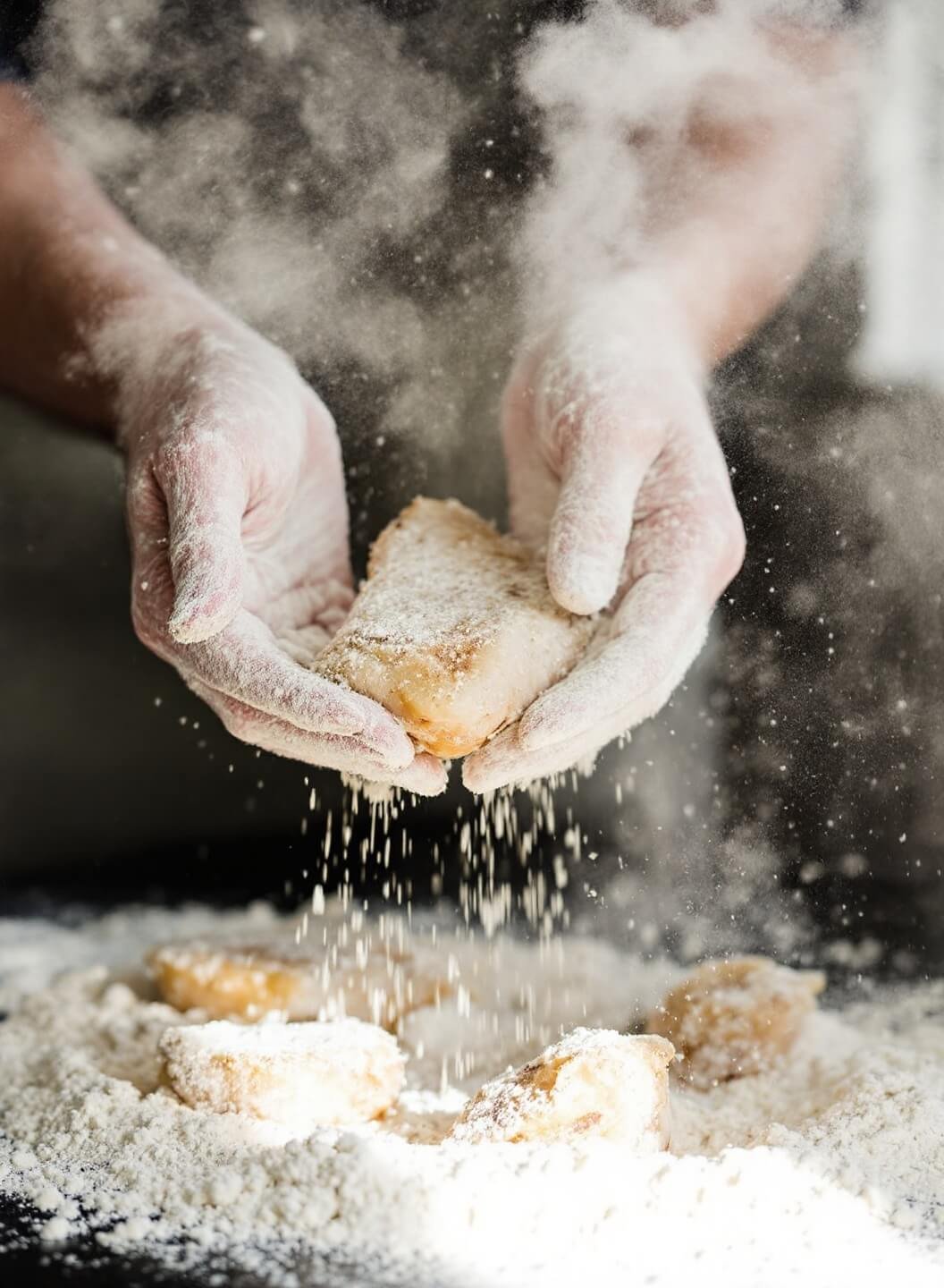 Hands coating a marinated chicken piece in seasoned flour under natural window light