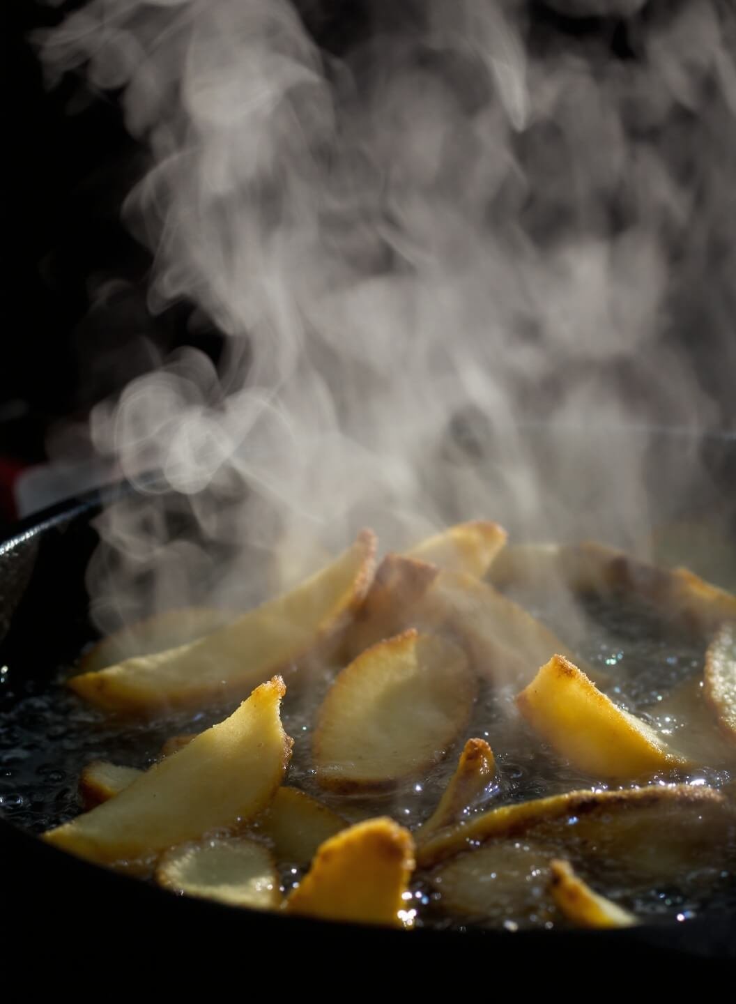 Deep frying chips in cast-iron pot with oil temperature at 350°F, dramatic lighting highlighting golden brown transformation and rising steam