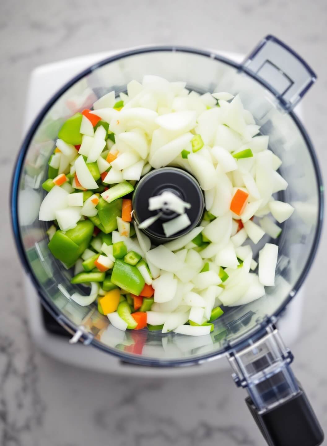 Close-up of a glass food processor bowl with diced onions and peppers, illuminated by overhead lighting.