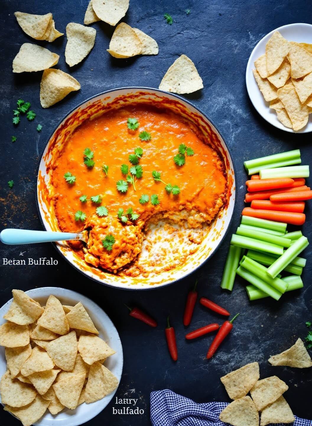 Hot buffalo dip in a baking dish with celery sticks, carrots, and tortilla chips on a dark stone surface