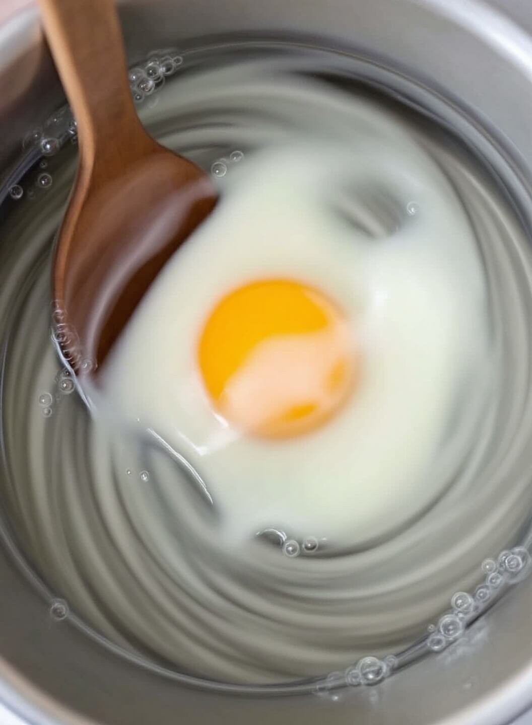 Perfectly formed poached egg in swirling water with rising bubbles and a wooden spoon stirring