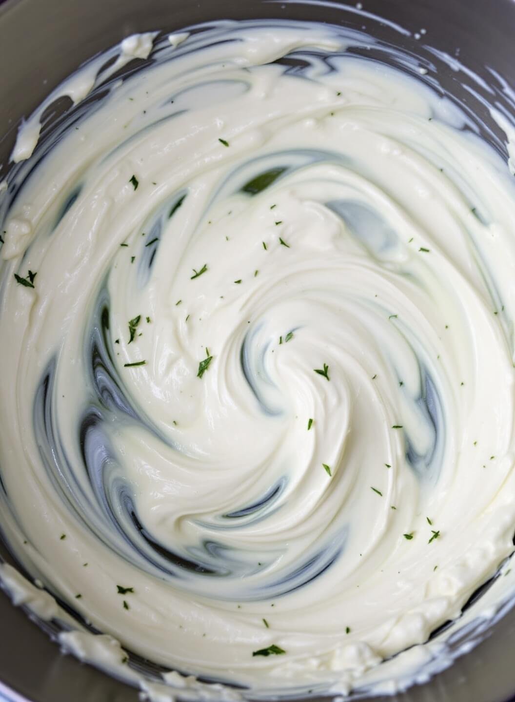 Velvety cream cheese and sour cream swirling together in a bowl under soft studio lighting, with scattered fresh herbs