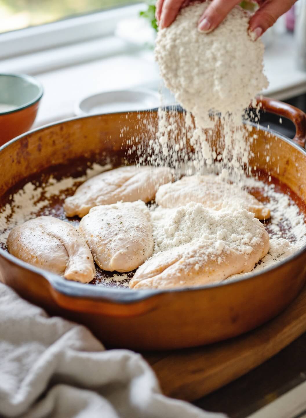 Hands coating seasoned chicken in flour in a rustic ceramic dish with natural window light in a kitchen setting