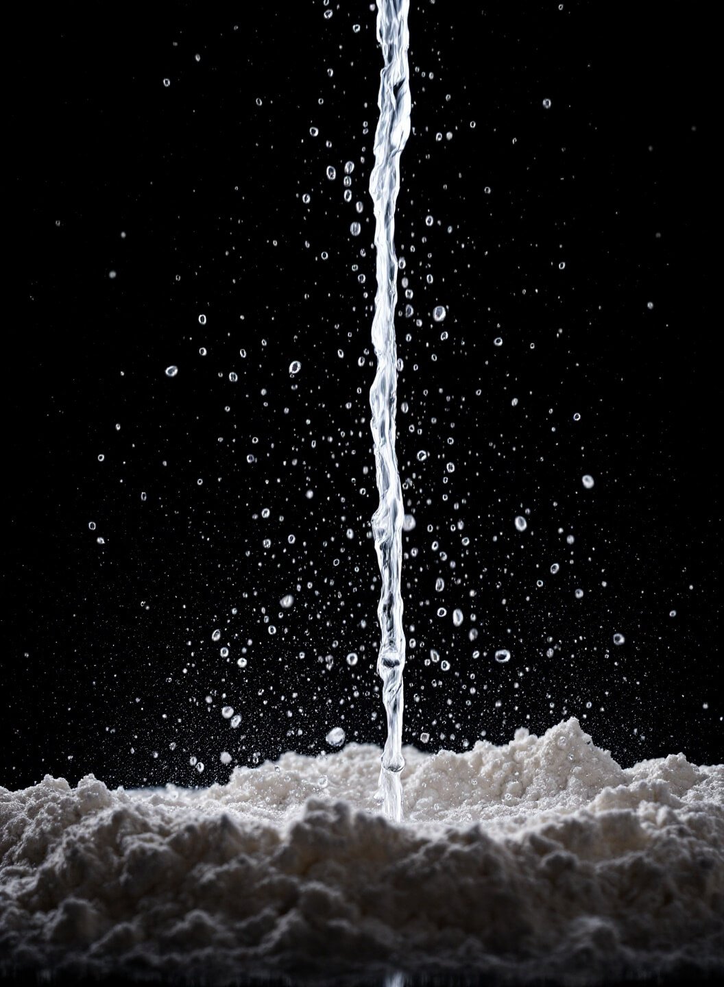 Dramatic image of ice water being drizzled onto flour mixture, with tiny water droplets illuminated against a dark background