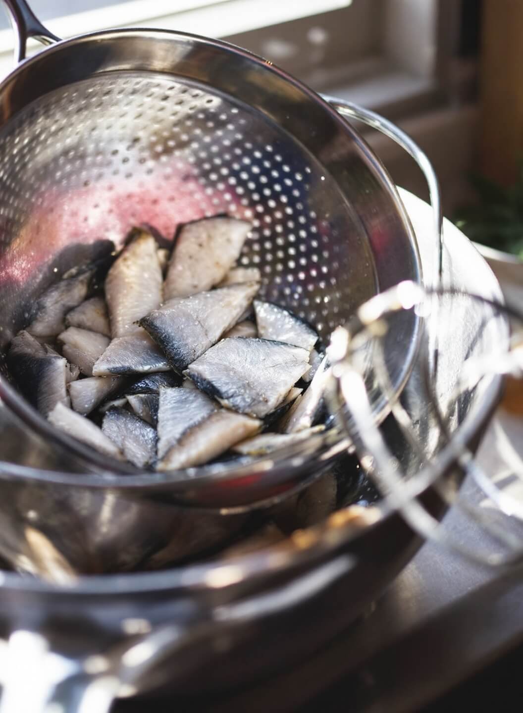 Mixing bowl with freshly drained and flaked tuna in a kitchen, stainless steel strainer visible, bathed in natural light from window, with water droplets