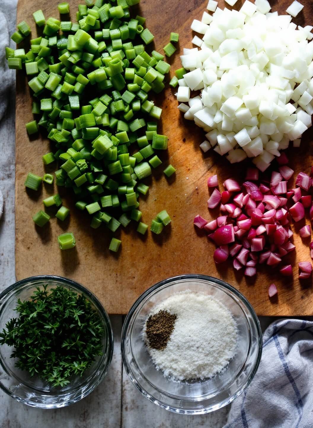 Diced celery and red onions with scattered herbs on a rustic wooden cutting board, crystal bowls containing seasonings nearby, all bathed in soft morning light.