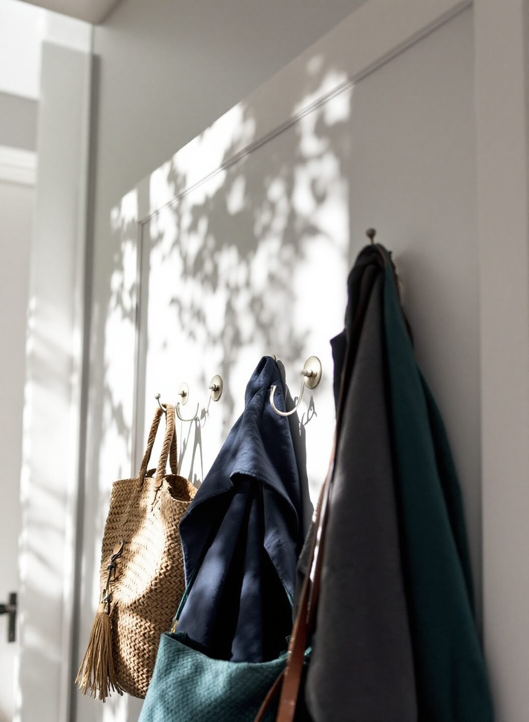 Low-angle view of a white door with over-the-door organization system of brushed nickel hooks holding various coats and bags, bathed in soft afternoon light, displaying a minimalist color palette with hints of navy and forest green.