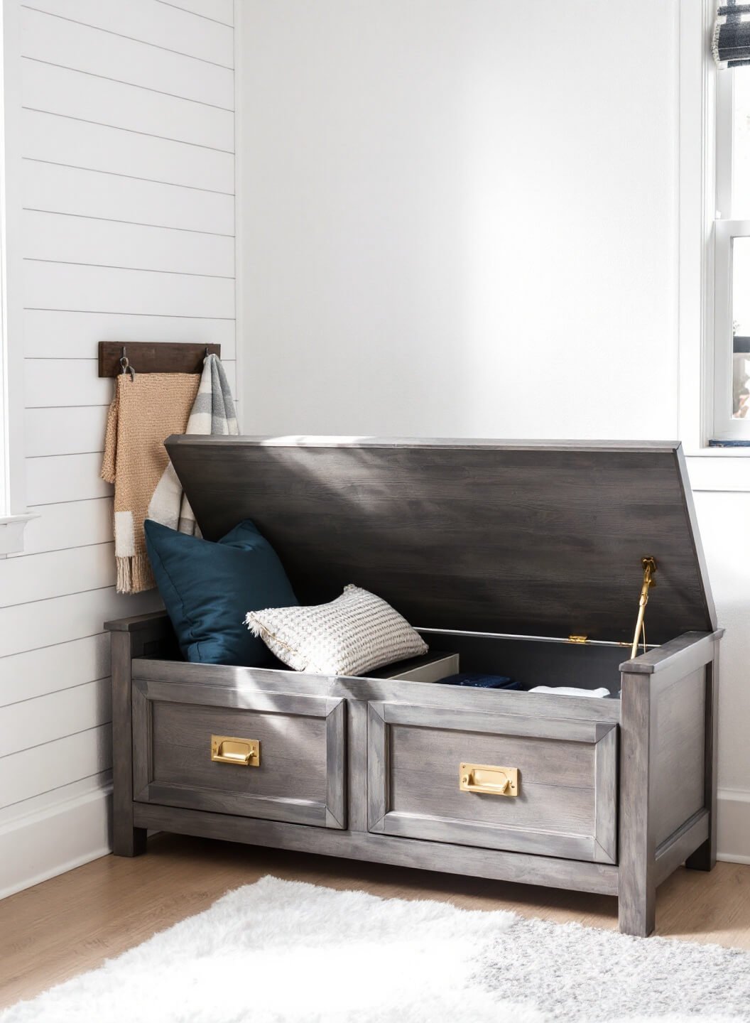 Multifunctional storage bench in weathered gray wood with brass hardware against a white shiplap wall, lid opened to reveal organized interior, adorned with navy cushion and textured throw pillows, in soft diffused daylight