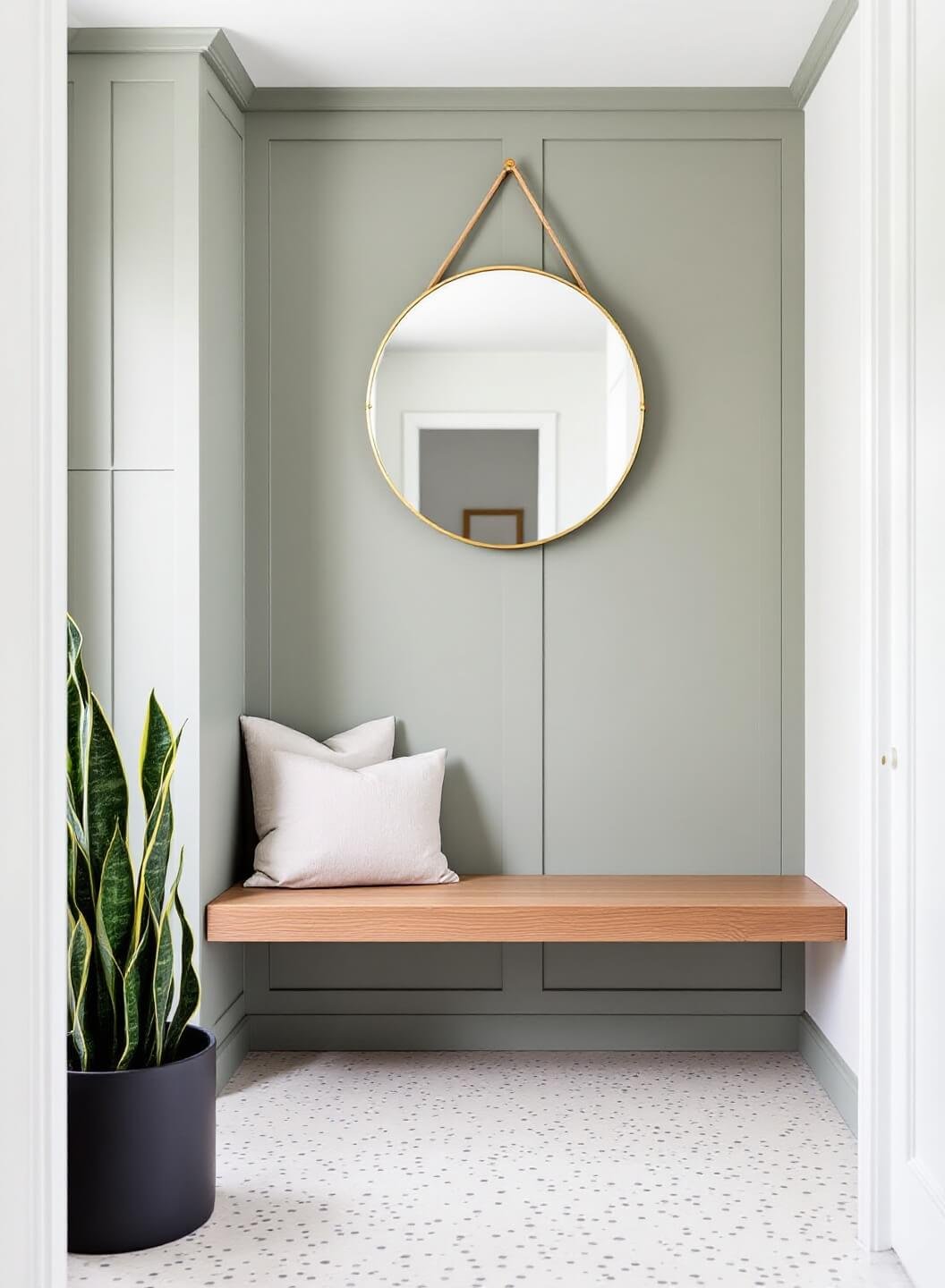 Contemporary Scandinavian style entryway with sage green built-in storage, brass mirror reflecting window light, floating oak bench with linen cushion, cream/grey terrazzo floor tiles, and snake plant in matte black planter