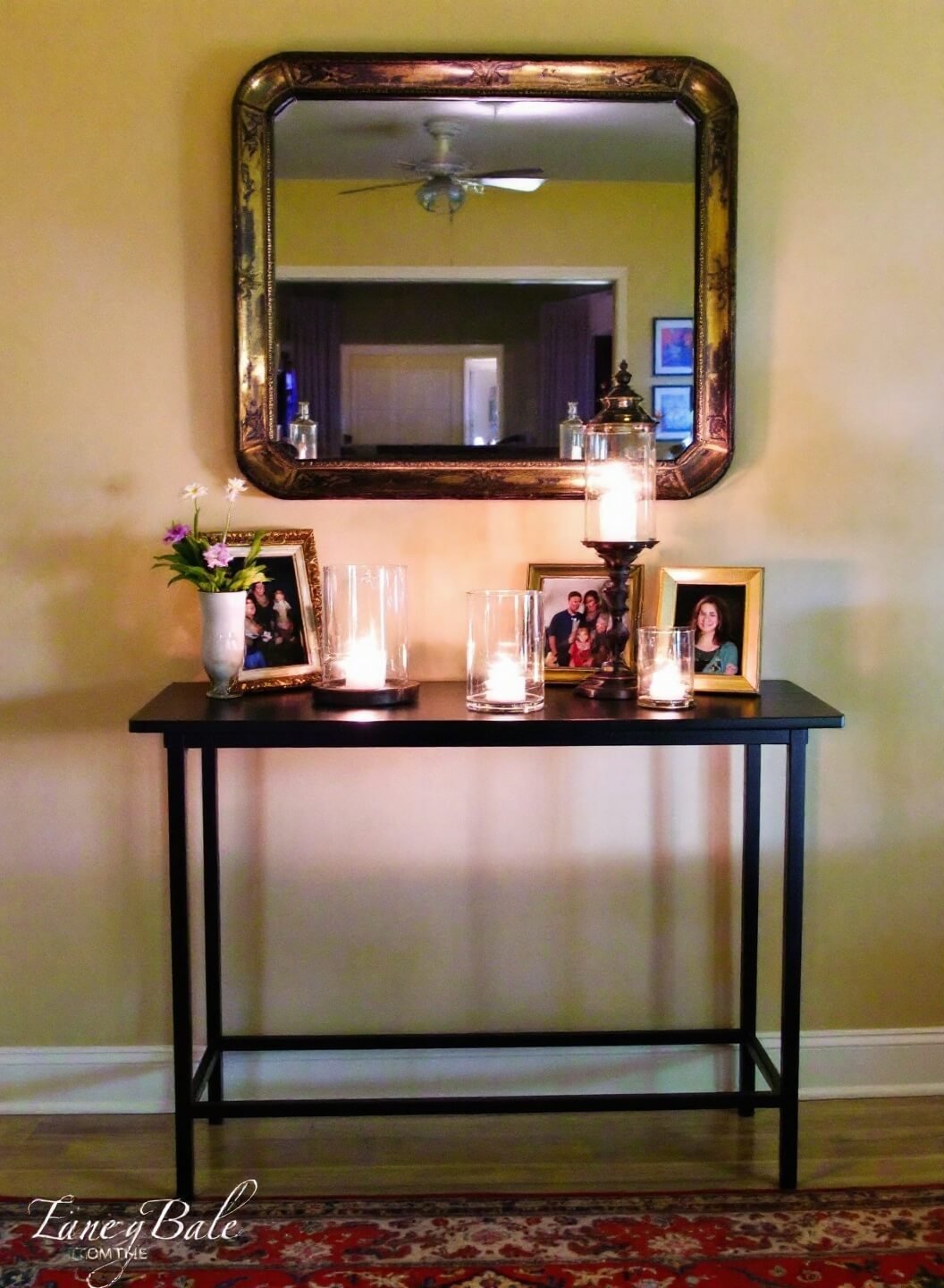 Antiqued mirror above a slim black console in a cozy evening entryway vignette with vintage Persian runner, glass hurricane lanterns with LED candles, and family photos in brass frames, reflected in mirror under moody lighting.