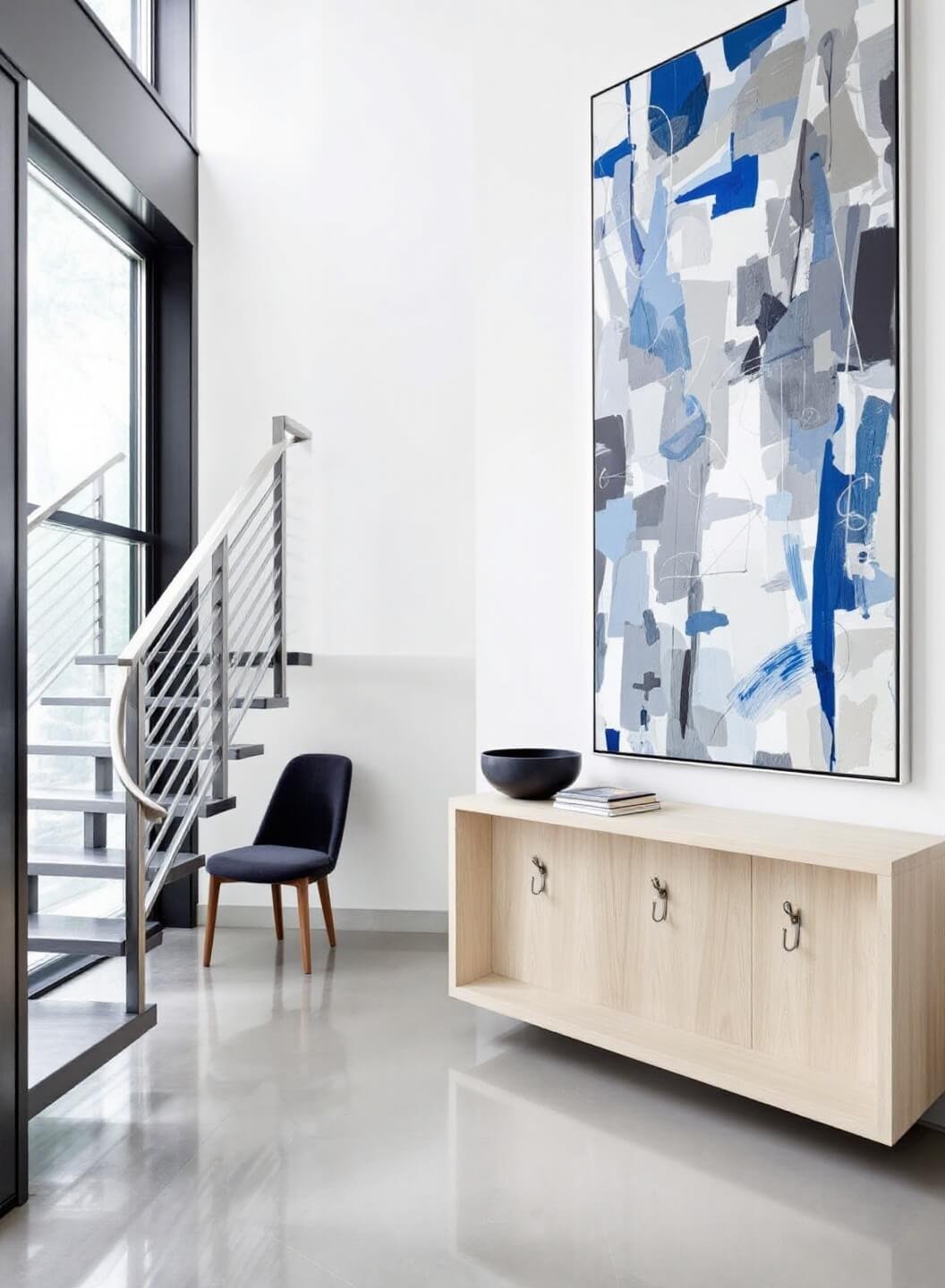 Contemporary foyer featuring floor-to-ceiling windows, a floating steel staircase, minimalist coat hooks, an oak console and abstract art, shot from the second floor highlighting geometric lines.