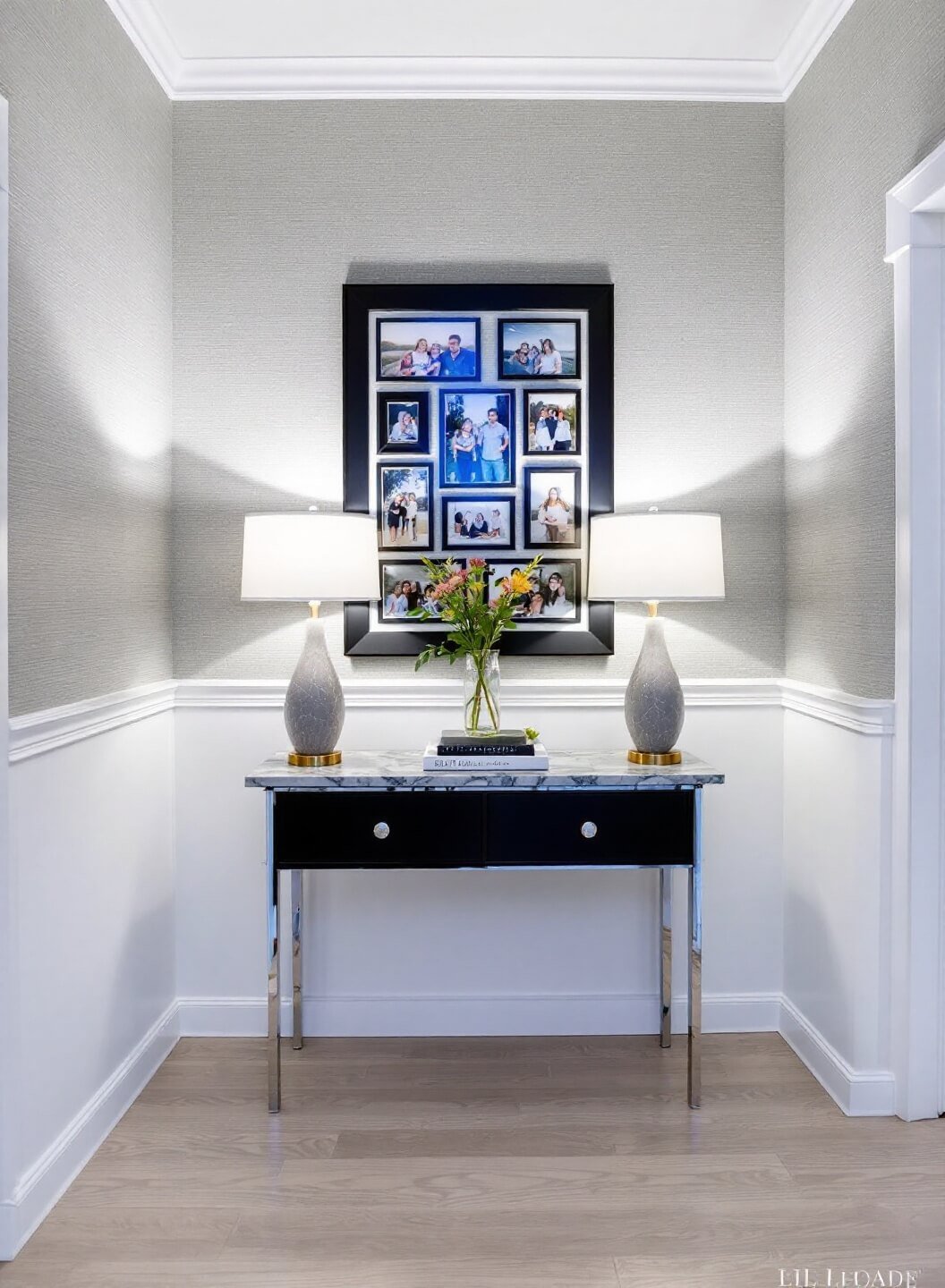 Transitional entryway with grey grasscloth wallpaper, white wainscoting, black-framed family photos, marble console with chrome legs, ceramic gourd lamps, under artificial and blue hour ambient lighting.