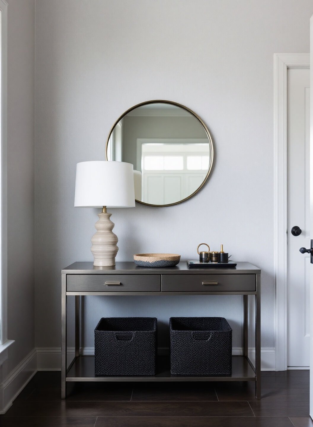 Contemporary foyer with a slim brushed steel console table, minimalist lamp and mirror, black water hyacinth baskets for storage, and a handmade ceramic catchall against a textured dove gray grasscloth wallpaper during blue hour.