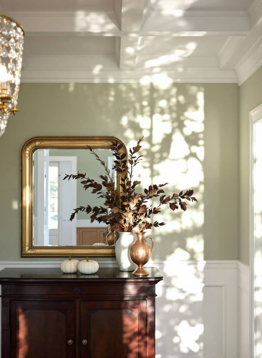 Traditional foyer with coffered ceiling, sage green walls, crystal chandelier, dark walnut console with brass-framed mirror, and autumn decorations in vintage copper vessels in afternoon light