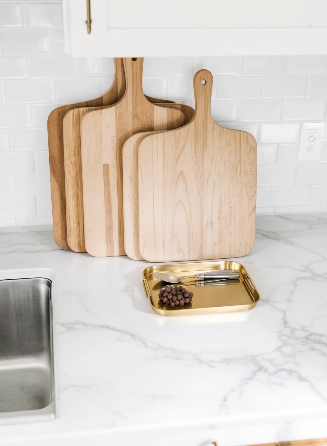 Overhead view of a minimalist prep zone with essential tools on a brass tray and wooden cutting boards against a subway-tiled backsplash on a white quartz counter, lit by natural light.