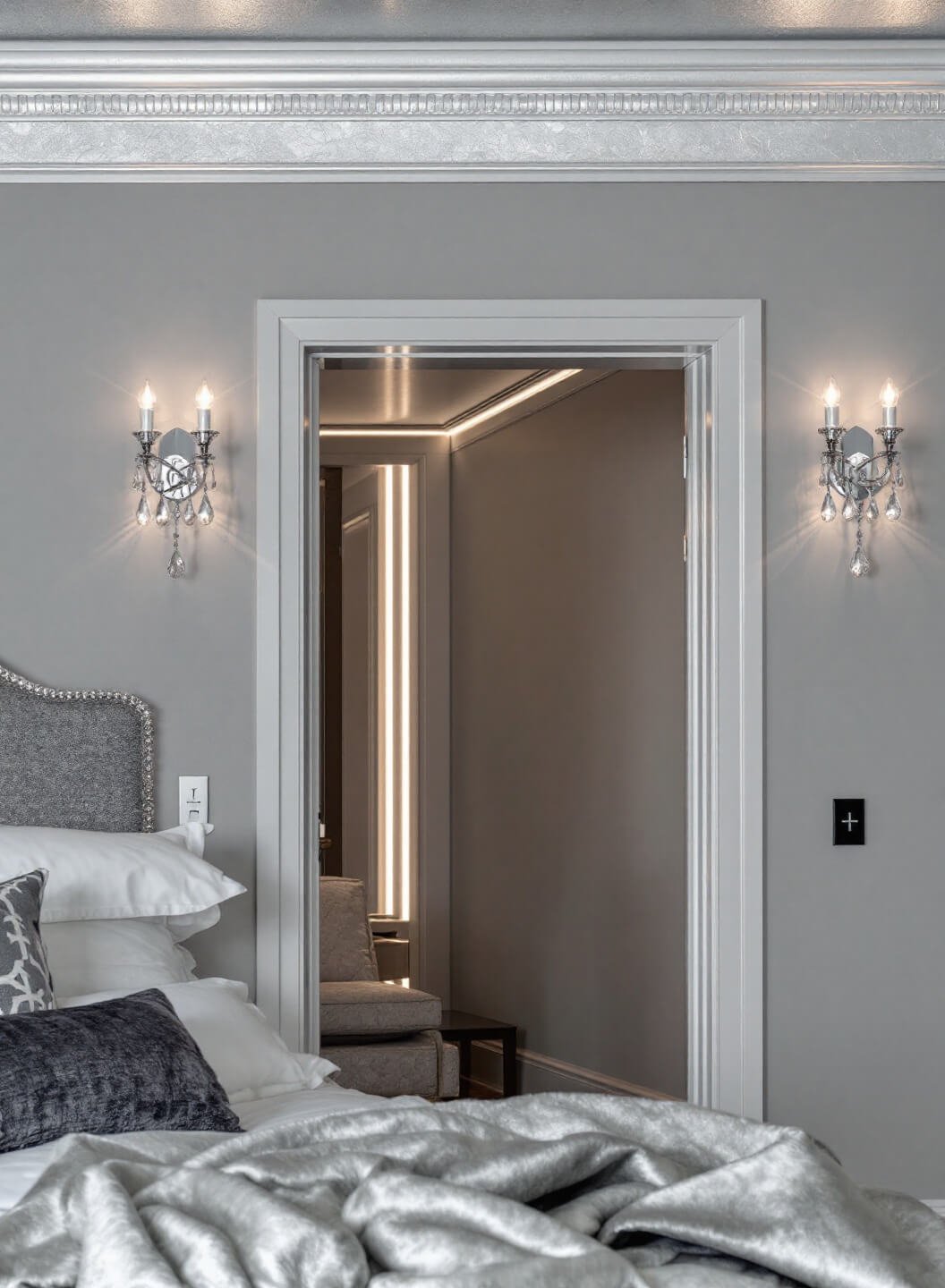 Elegant evening bedroom scene showing cool grey walls, silver leaf ceiling detail, twin crystal sconces, and LED strips behind an upholstered headboard with textural velvet, silk, and brushed metal finishes