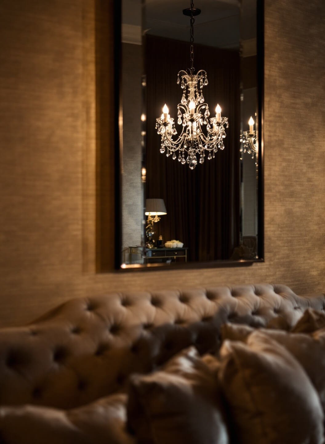 Boutique-style bedroom with warm beige grasscloth wallpaper, tufted headboard, crystal chandelier, and strategic mirror placement during dusk